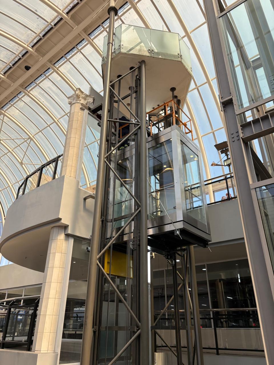 Glass passenger-elevator cabin within a steel structure in a glazed atrium.